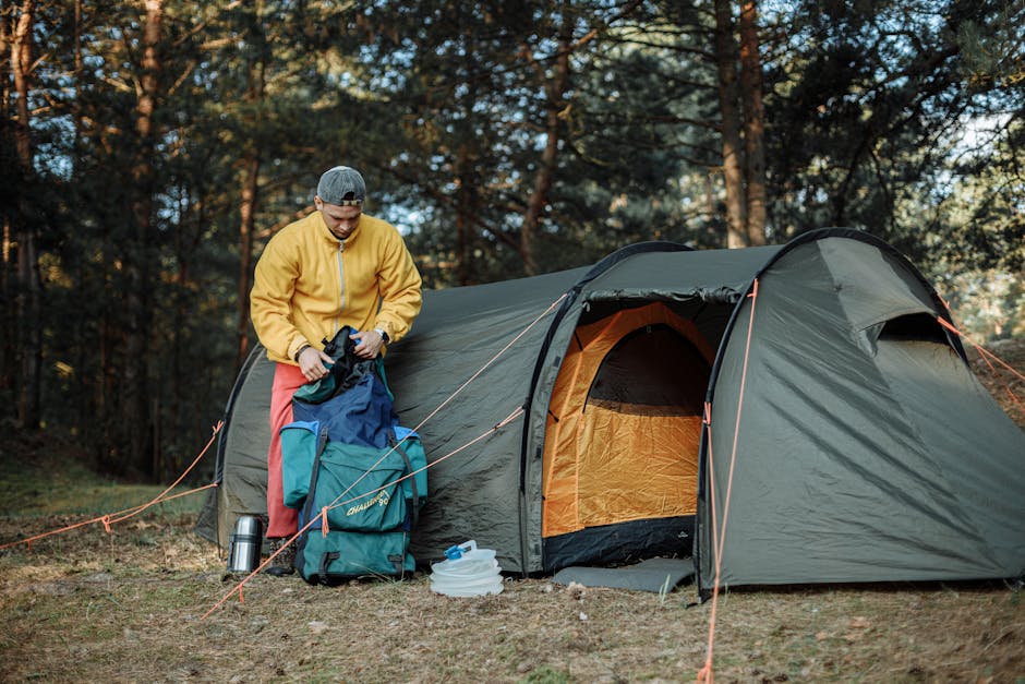 A young man organizing camping gear beside a tent in a scenic forest.