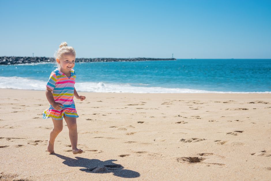 A happy little girl with blond hair runs on a sunny beach with the sea in the background.