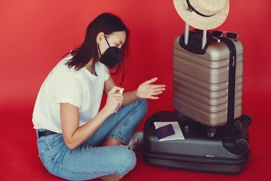 Side view of young woman in casual clothes and medical mask sitting near luggage with passport and tickets while disinfecting hands on red background