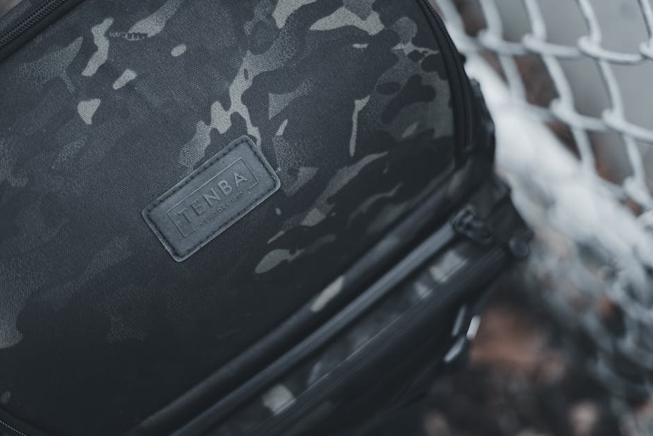 Camouflage Tenba backpack close-up near chain-link fence, highlighting the brand logo.