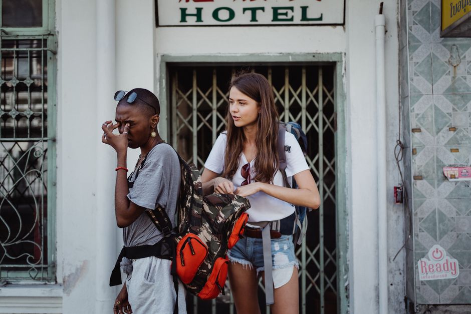 Two travelers with backpacks take a break outside a hotel, showing expressions of fatigue and discussion.