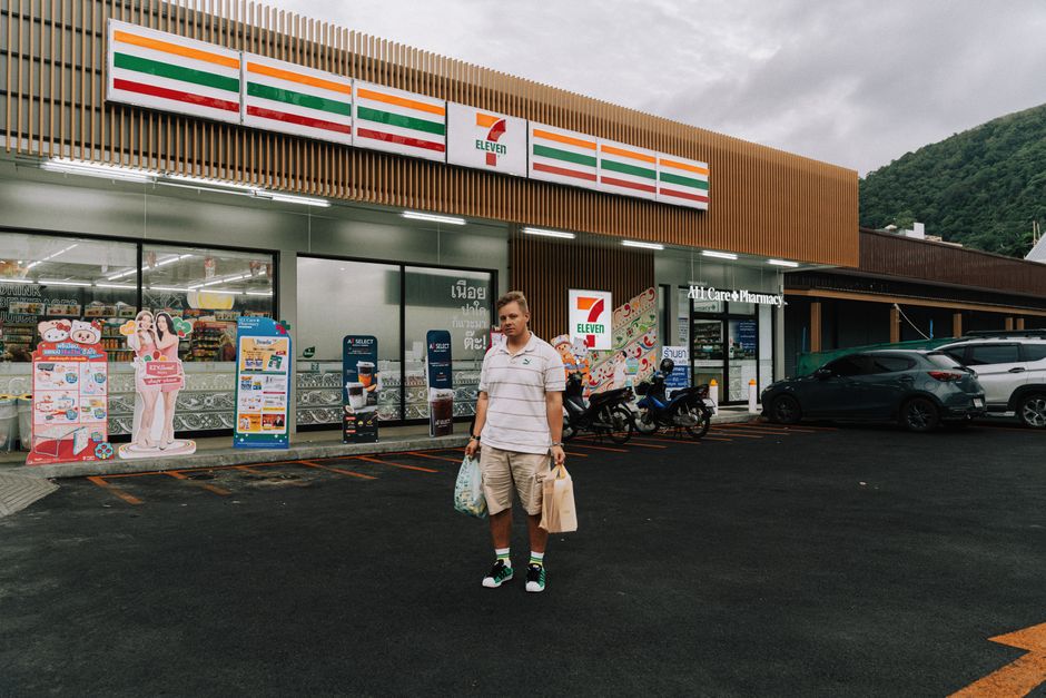 A man stands outside a 7-Eleven store in Phuket, Thailand, holding shopping bags.