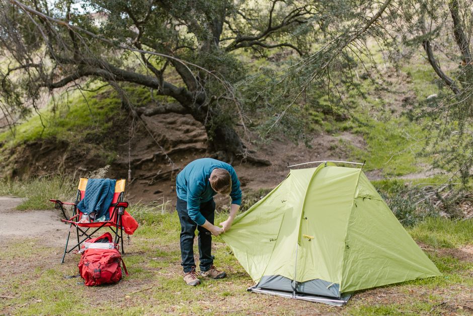 A man in blue sets up a green tent in a peaceful outdoor setting with a chair and backpack.
