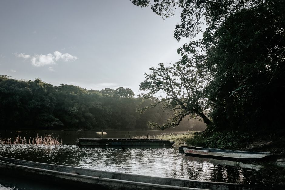 Serene lake scene with wooden boats, lush trees, and a beautiful sky at sunrise.