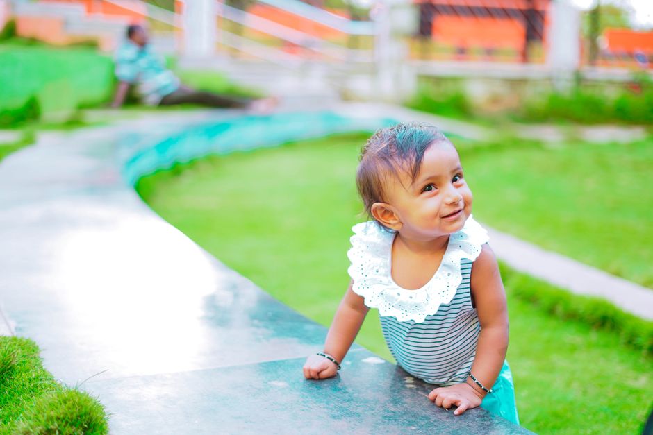 High angle of adorable toddler girl standing on green lawn of playground leaning on hands and smiling