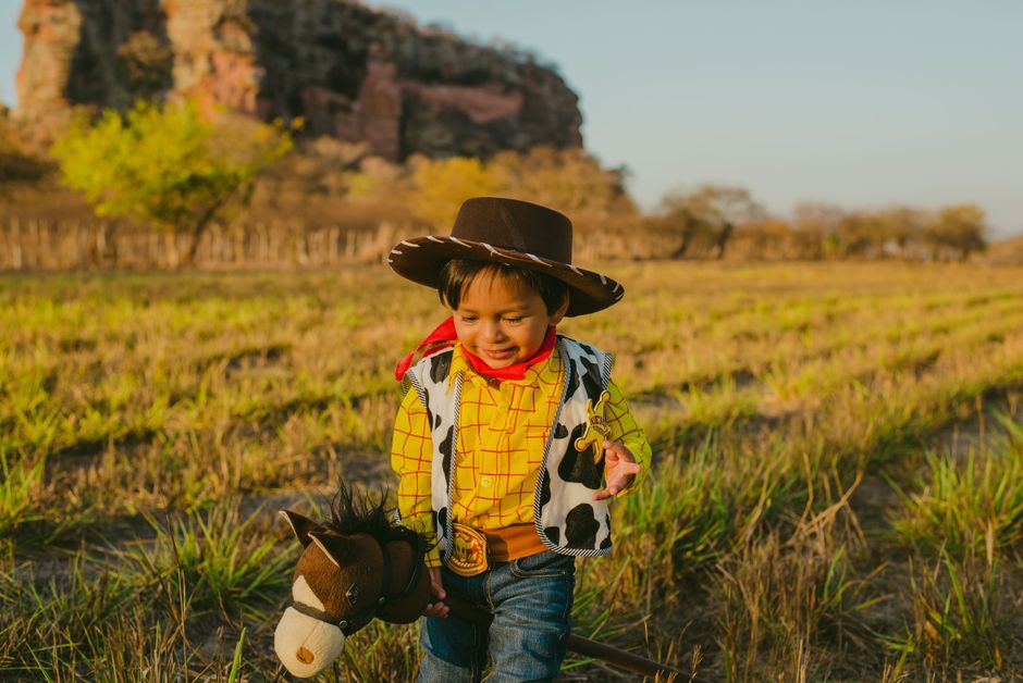 A young boy in a cowboy costume joyfully plays outdoors with a toy horse.