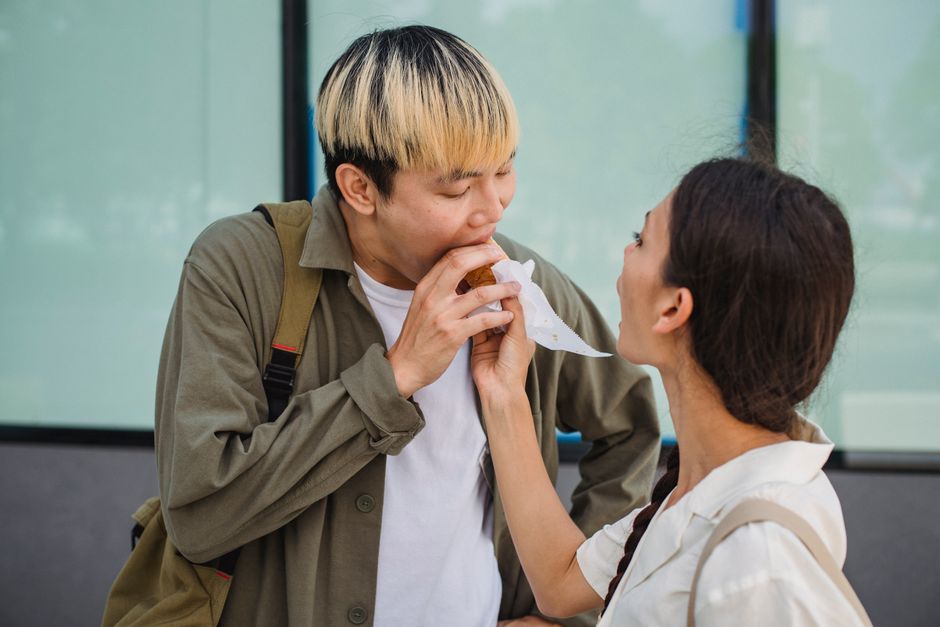 Cheerful young Asian female hand feeding hungry positive boyfriend with delicious hot dog while standing together on street