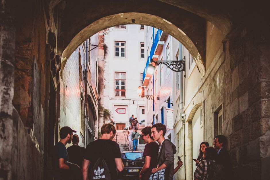 A group of people socializing in a historic Lisbon alleyway, under a stone archway, during daytime.