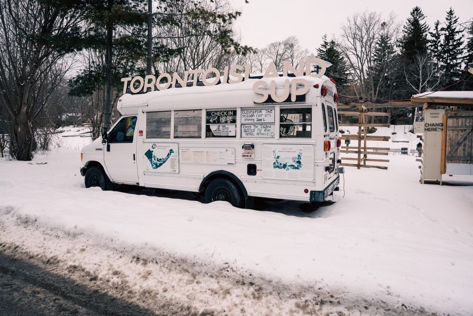 A snow-covered bus on Toronto Island during winter, with signs and bare trees in the background.