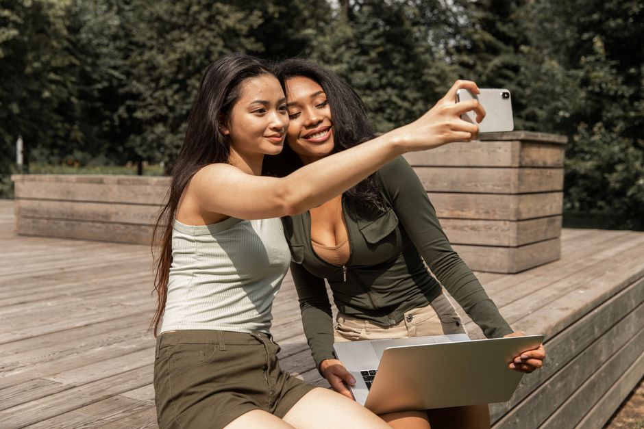 Positive young multiethnic ladies with long dark hairs in casual outfits smiling and taking selfie on mobile phone while chilling in park with laptop