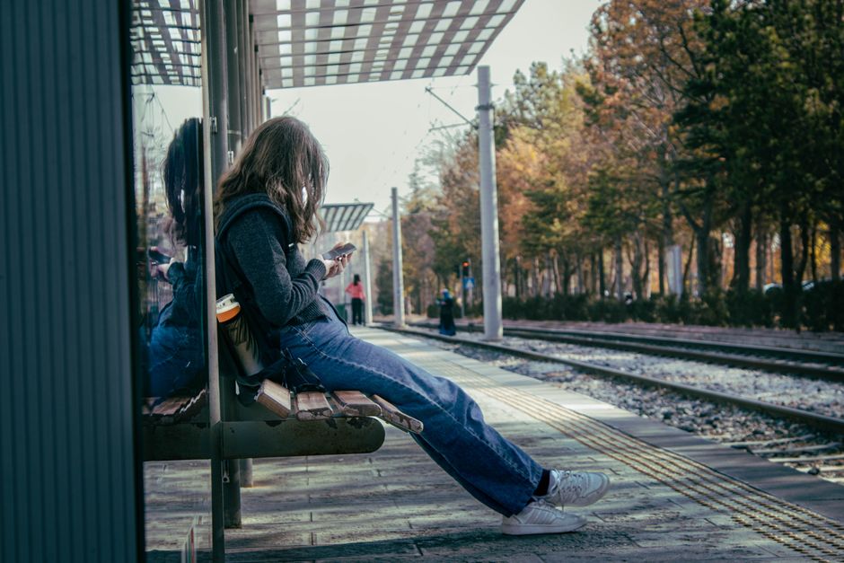 A young woman using a smartphone while waiting at a tram station in autumn.