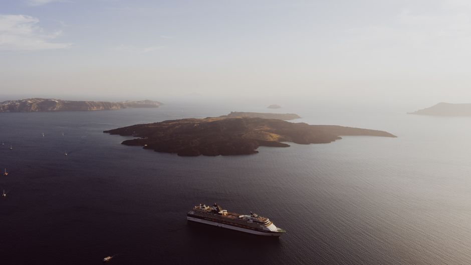 Aerial photo of a cruise ship sailing near Santorini island under a serene sunset sky.
