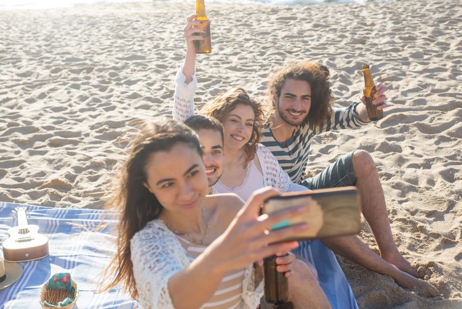 Group of young friends taking a selfie during a sunny beach picnic, smiling and enjoying drinks.