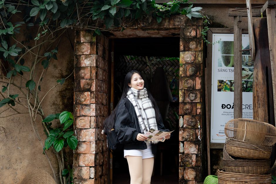 A woman stands at a rustic Vietnamese entrance, holding a map and smiling.