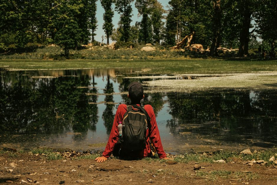 A teenager with a backpack relaxes by a reflective lake on a warm summer day, surrounded by trees.