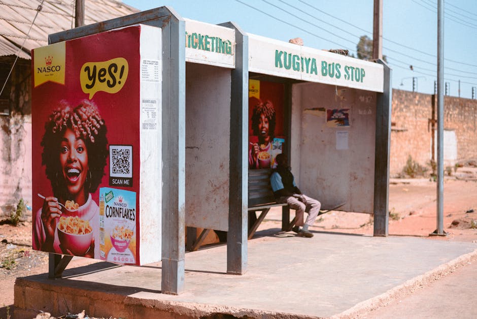 A sunny day at Kugiya Bus Stop in Jos, featuring vibrant advertising and urban life.