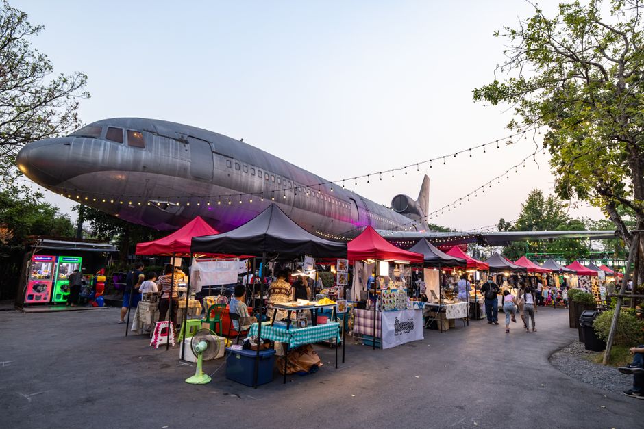 Bustling outdoor market scene with diverse stalls under an airplane, vibrant lights and evening atmosphere.