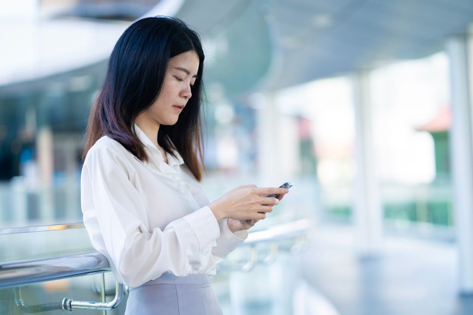 Confident Asian woman in smart casual attire uses smartphone, indoors.